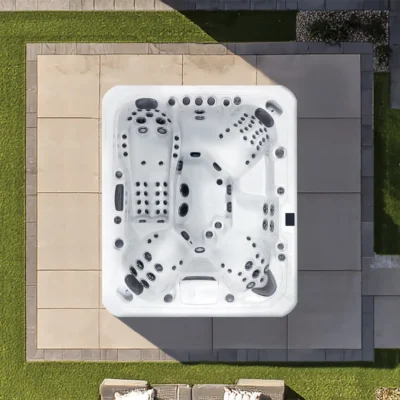 Top-down view of a white outdoor hot tub with many jets on a beige tile patio, surrounded by green lawn.