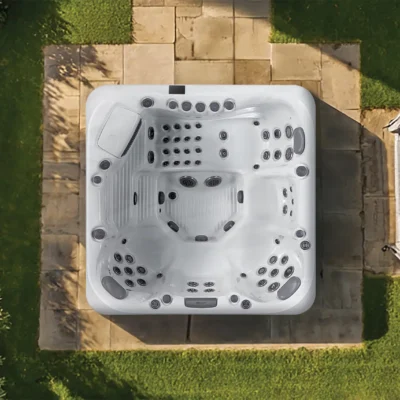 Top-down aerial view of a white square hot tub with multiple jet nozzles and built‑in seating on a stone patio, surrounded by grass and hedges to the left and right.