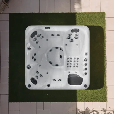 Top-down view of a white outdoor hot tub with numerous jets on a tiled patio bordered by artificial green grass and plants nearby