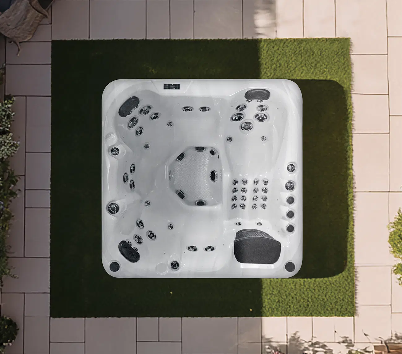 Top-down view of a white outdoor hot tub with numerous jets on a tiled patio bordered by artificial green grass and plants nearby