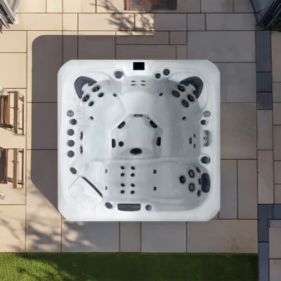 Top-down view of a white square hot tub with numerous jets, installed on a tiled patio beside a green lawn area.