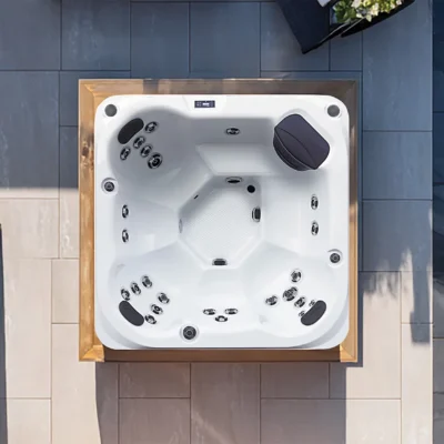 Top-down view of a white outdoor hot tub with multiple jets and a dark headrest, set in a wooden frame on a tiled patio.