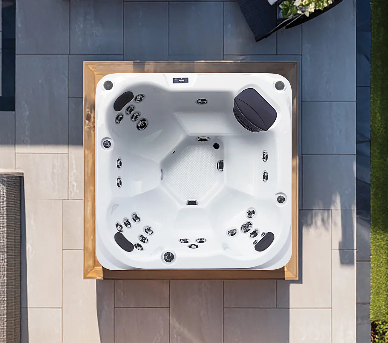 Top-down view of a white outdoor hot tub with multiple jets and a dark headrest, set in a wooden frame on a tiled patio.